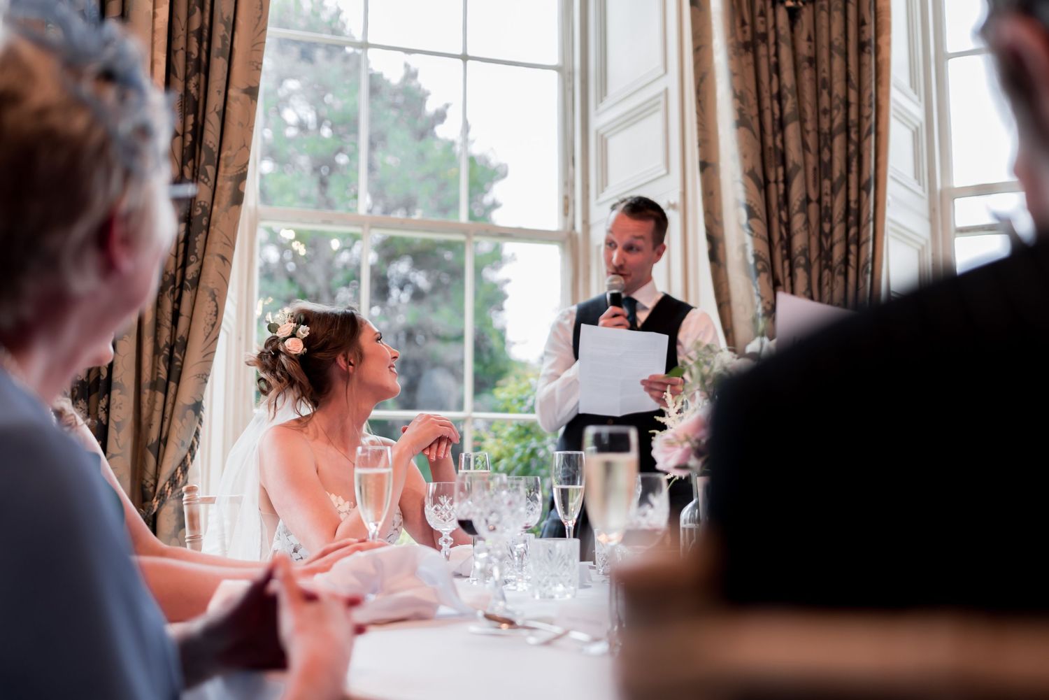 A waiter takes an order at an elegant dinner party while guests sit at a table by tall curtained windows.
