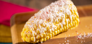 A grilled corn on the cob topped with grated cheese and spices sits on a wooden cutting board against a red background.