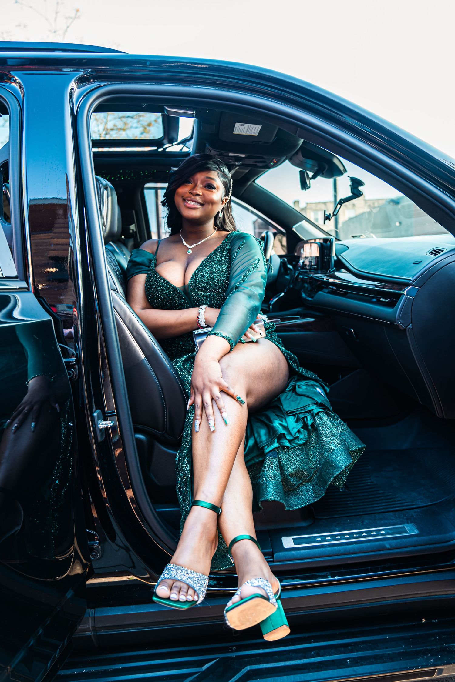 Someone sits in a luxury car wearing a sparkly emerald green dress and silver sandals with a casual, relaxed pose.