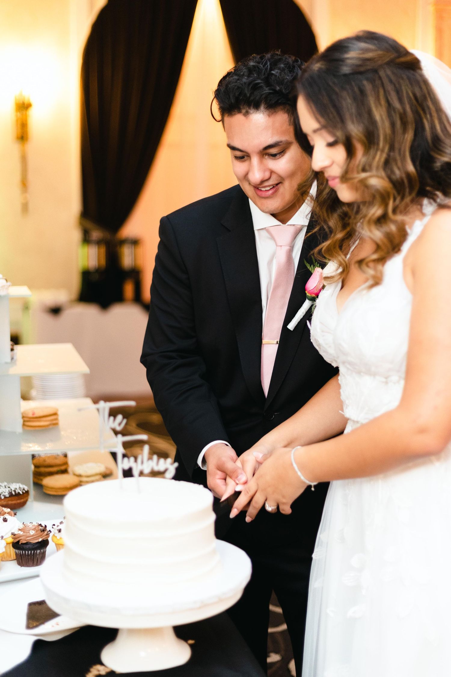 A bride and groom cut their wedding cake together at an elegant reception with cupcakes and desserts displayed.