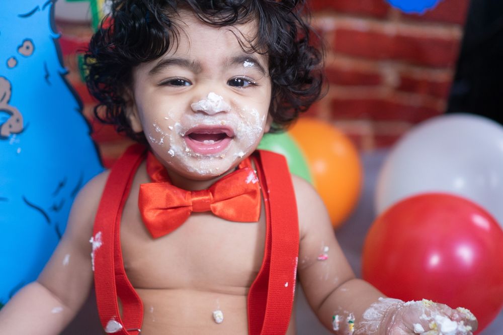 Toddler in red bow tie with messy face enjoying celebration with balloons.