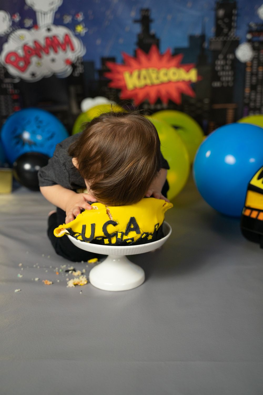 Yellow cake on white pedestal with comic book style backdrop and colorful balloons.