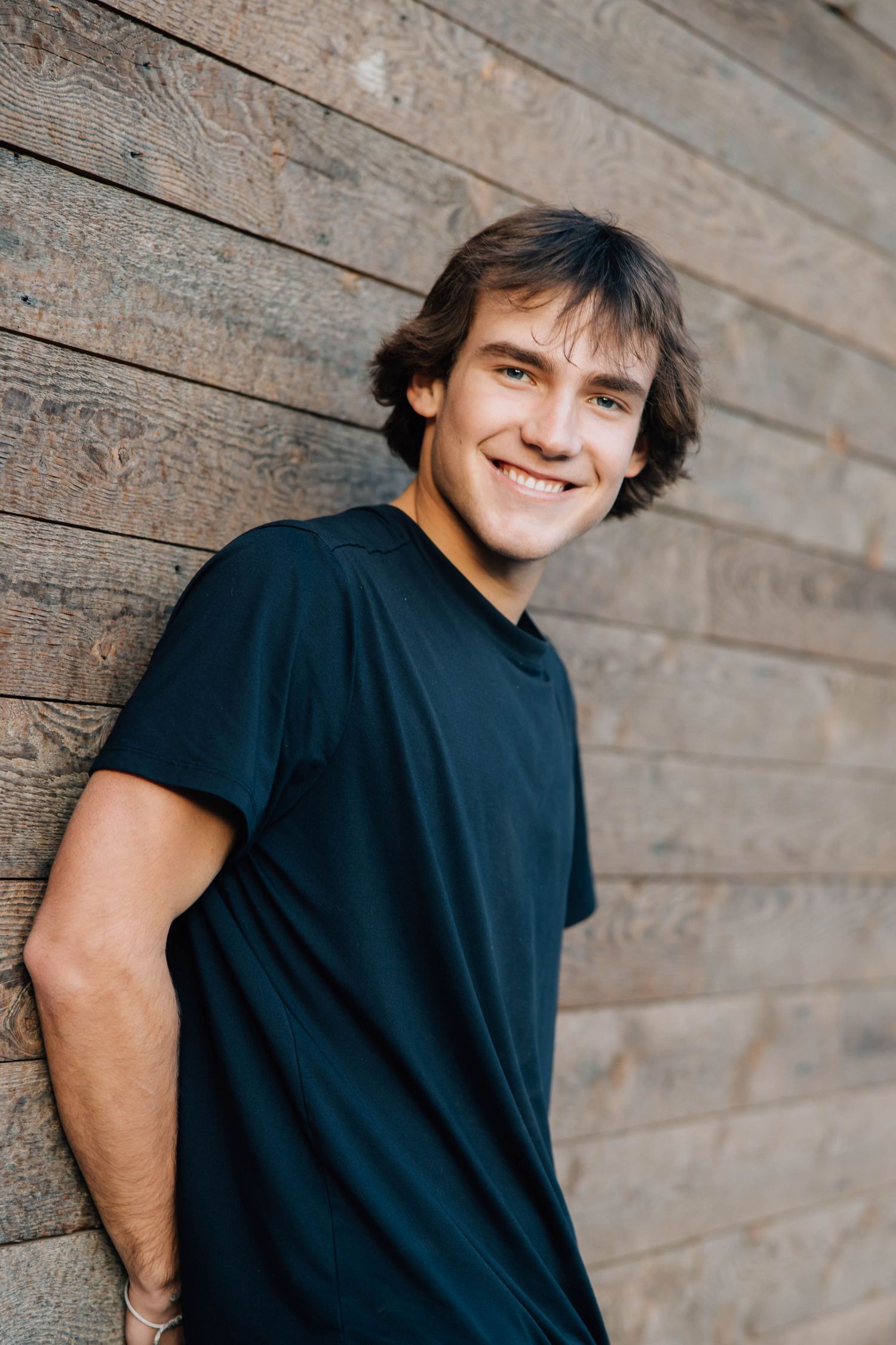 Smiling young man with brown hair wearing a black t-shirt leaning against a rustic wooden wall.