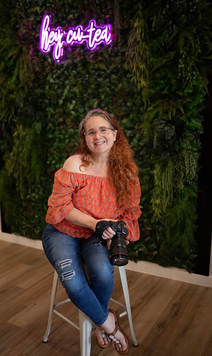 Professional photographer sits on stool with camera against green foliage wall with pink neon 'by a tea' sign.