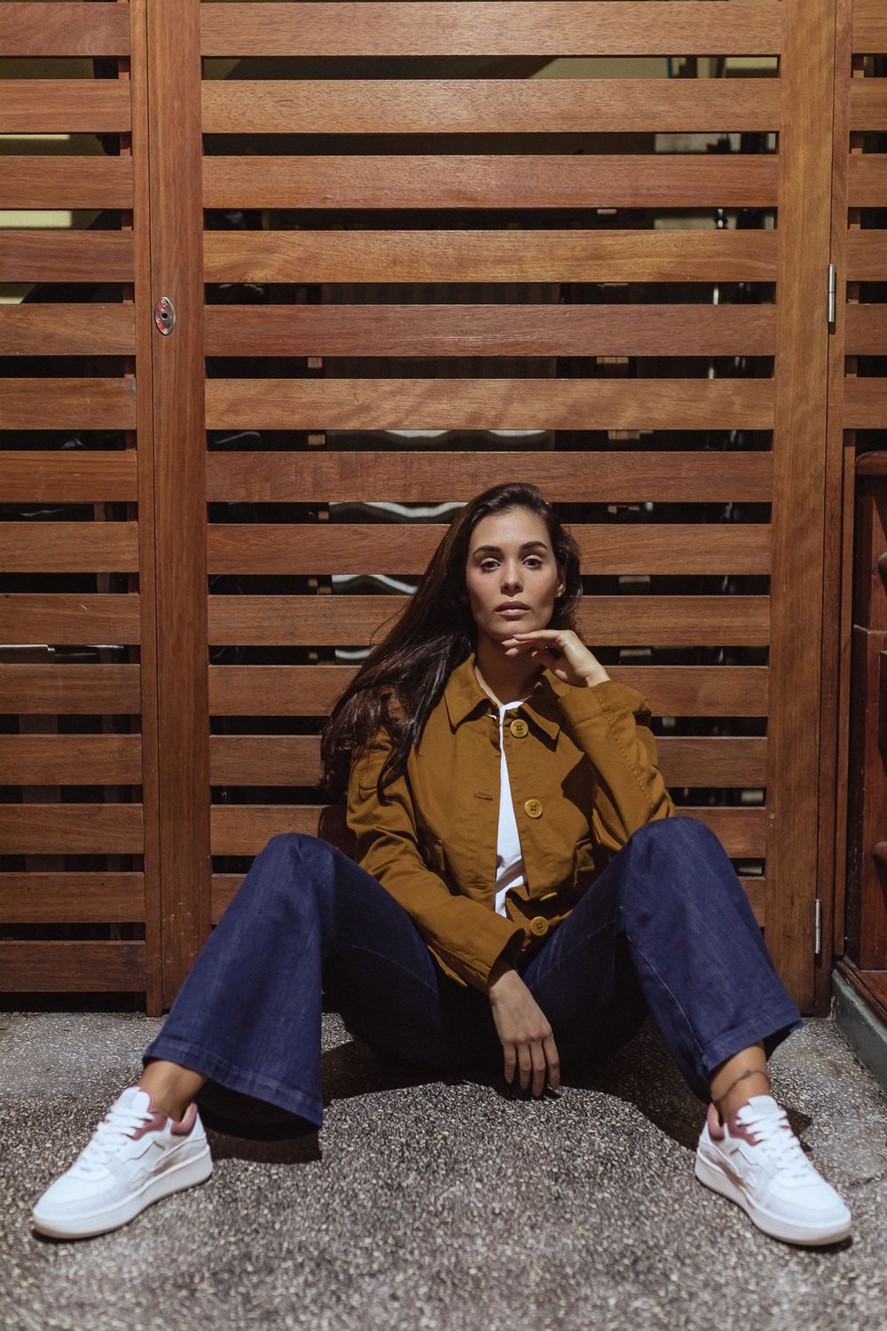 A casual streetwear look featuring wide-leg jeans, white sneakers and a brown jacket against a wooden slatted backdrop.