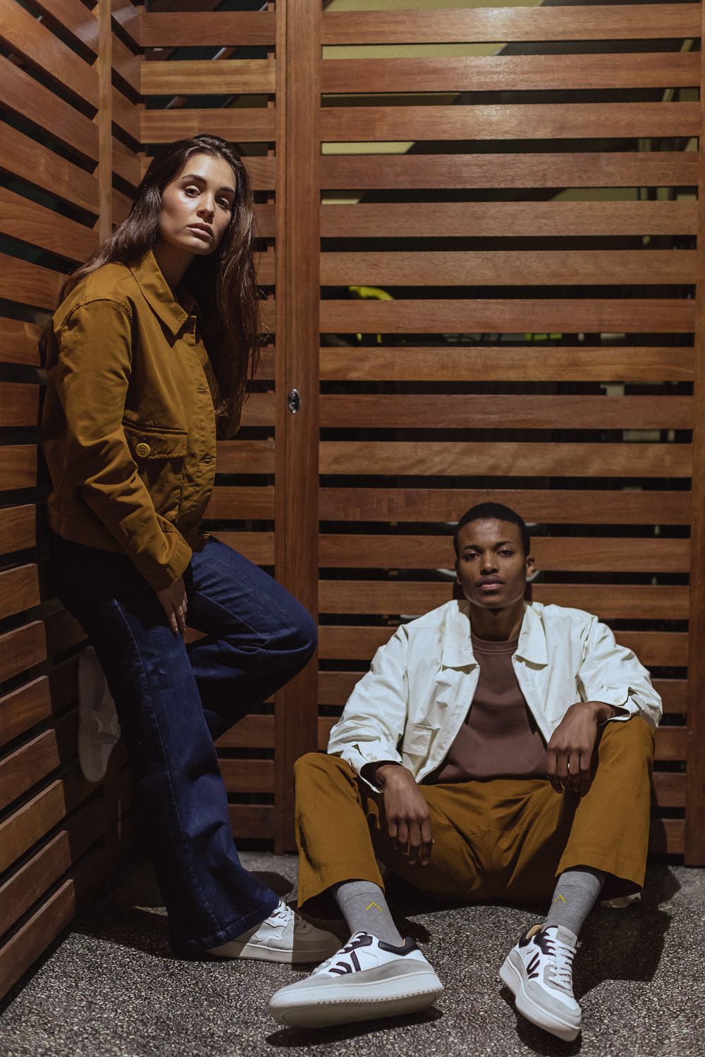 Two models in casual street style clothing pose against wooden slat wall wearing mustard jacket and white denim outfit.