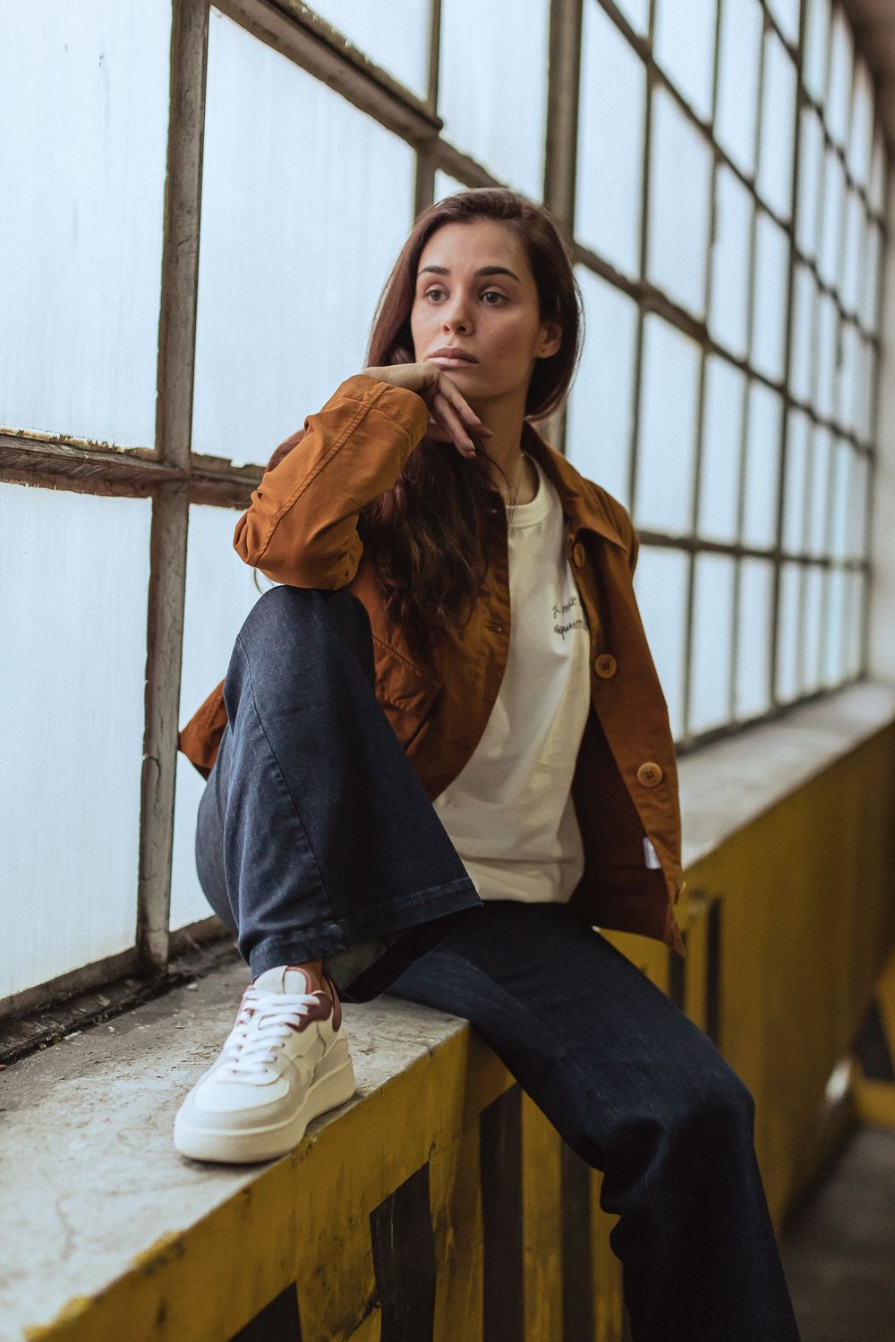 Casual streetwear look featuring brown jacket, white sneakers and dark pants while sitting on windowsill in industrial setting.