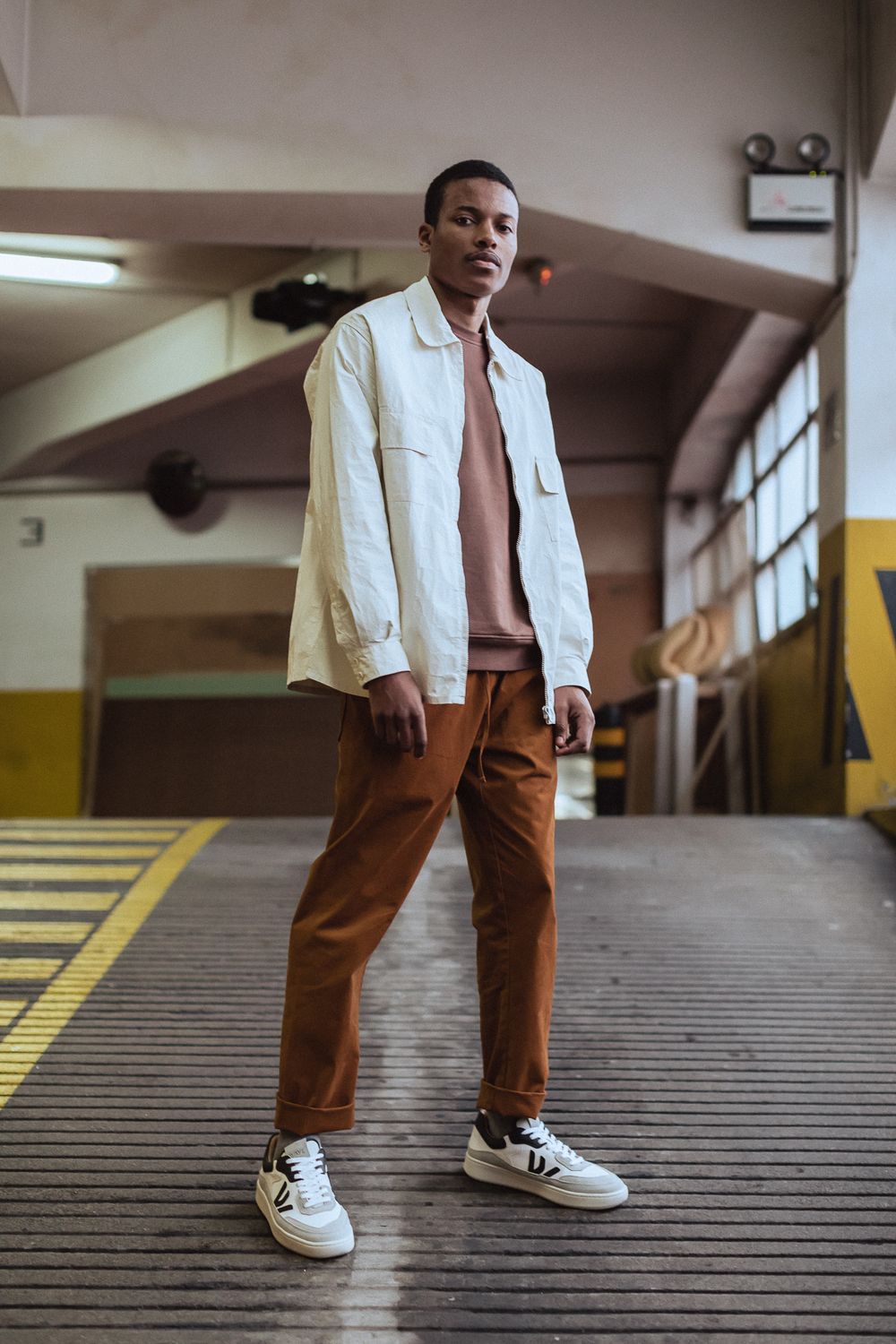 Stylish casual outfit featuring brown pants, white jacket and sneakers photographed in an urban parking garage setting.