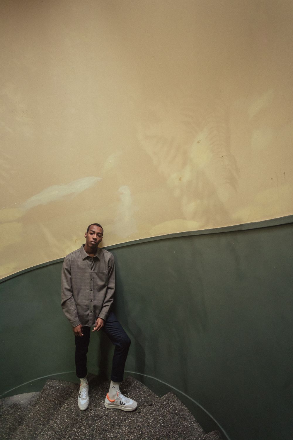 Figure stands against two-toned wall wearing gray button-up shirt, dark pants and white sneakers in minimalist setting.