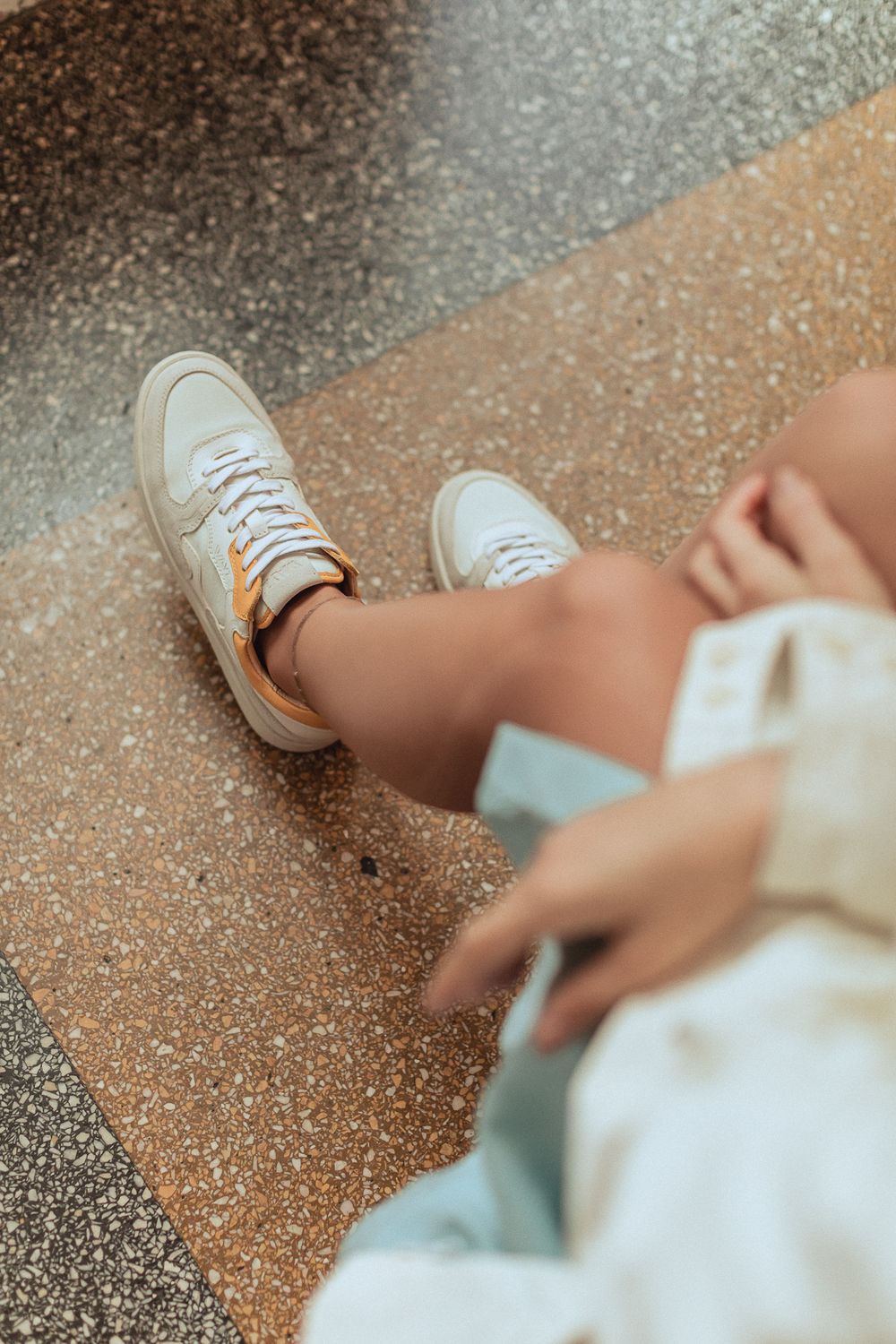 Person wearing white sneakers and light colored clothing sits on textured ground in artistic lifestyle composition.