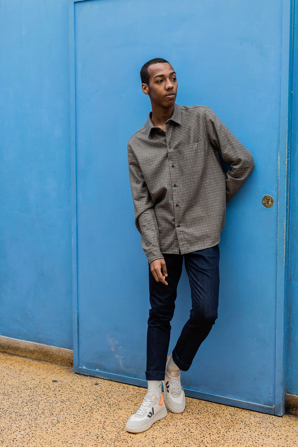 Person in grey button-up shirt and dark jeans poses casually against bright blue door wearing white sneakers.