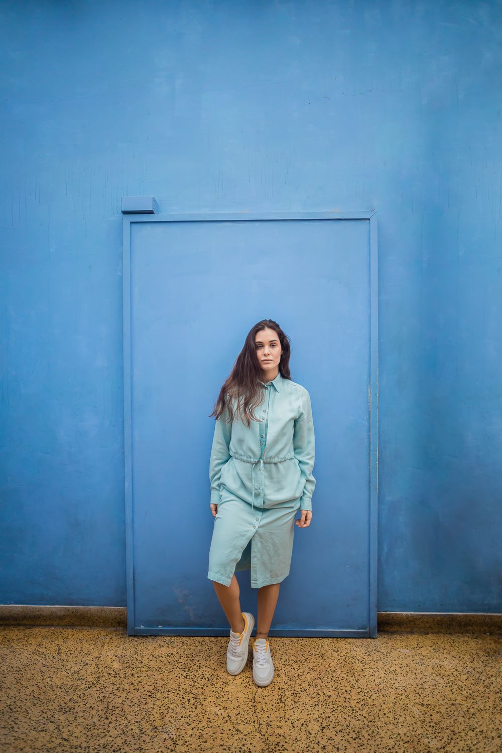 Figure in a light blue shirt dress poses against a matching blue industrial door with textured floor beneath.