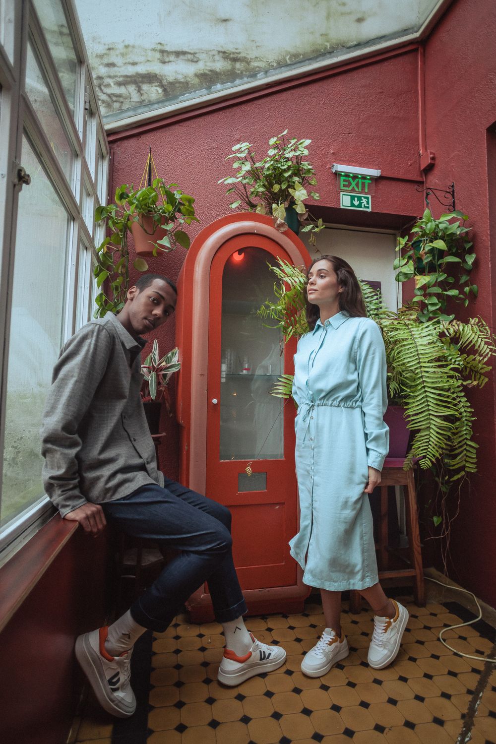 Two people stand together in front of a red door with plants and ferns hanging in a tiled entryway.