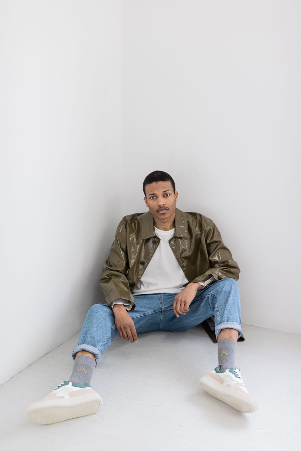 Person wearing olive jacket, white t-shirt, cuffed jeans and white sneakers sits casually against white walls.
