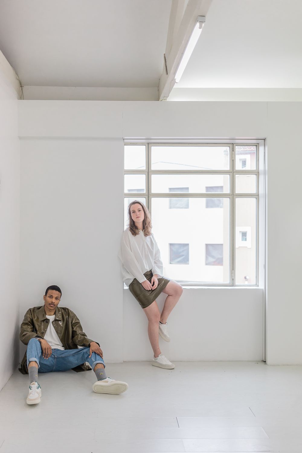 Two people relaxing in a minimalist white room with one sitting on windowsill and another on floor.