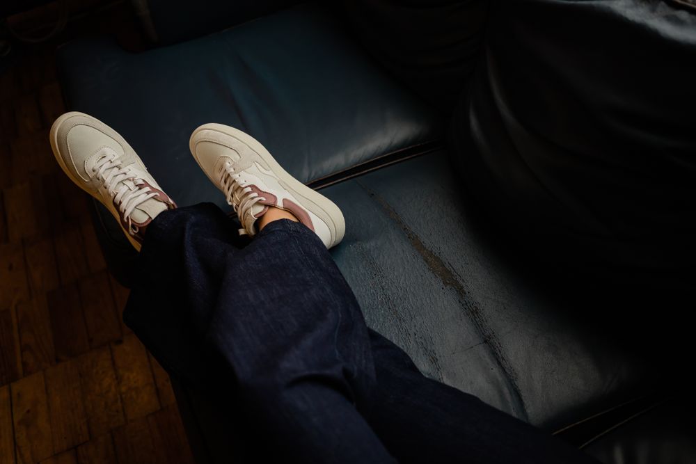 Person wearing blue jeans and white casual sneakers sitting on dark leather couch against wooden floor.