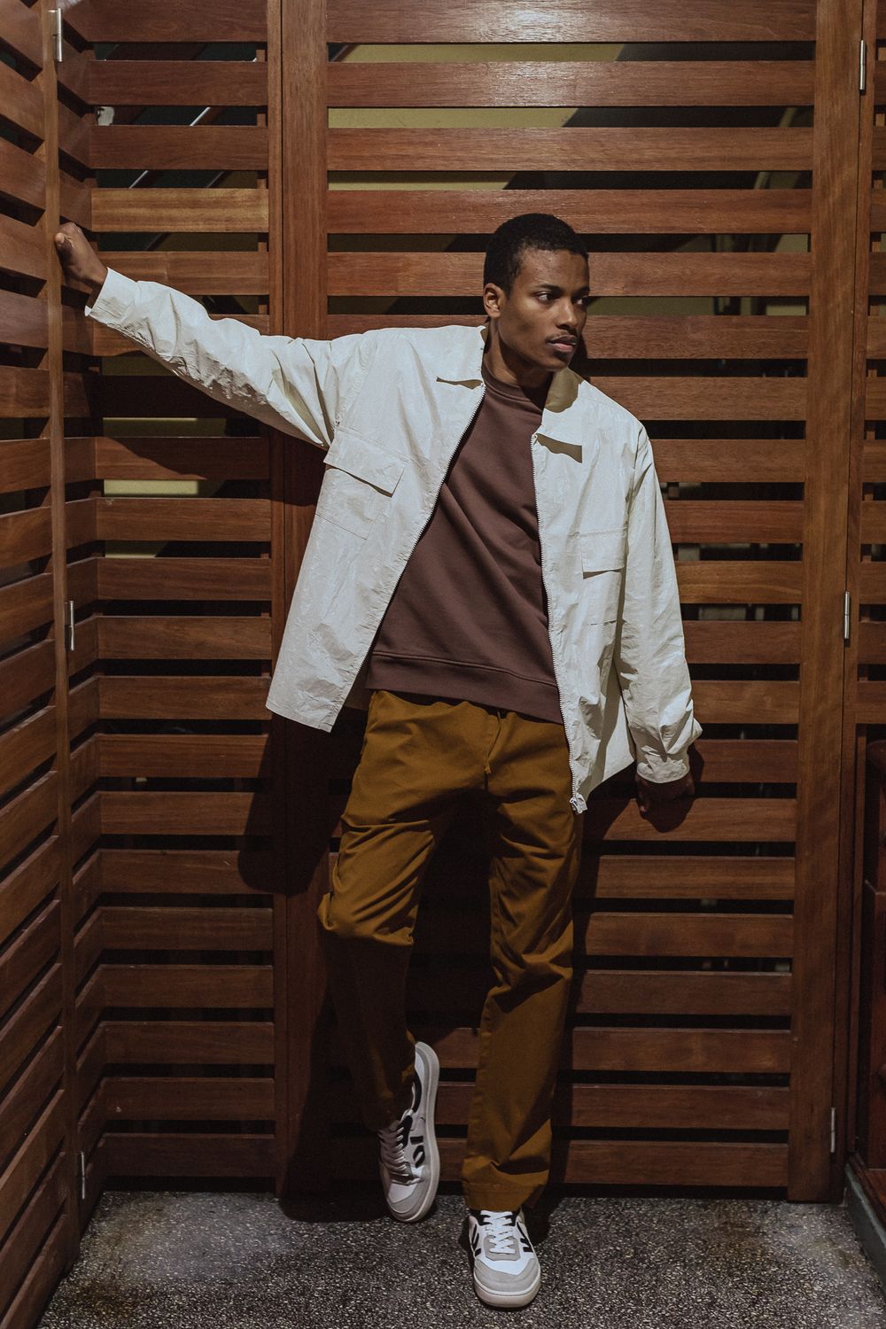 A person in a white jacket and brown pants poses against wooden slats wearing white sneakers.