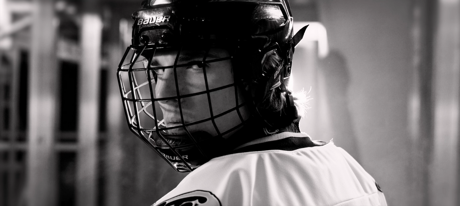 A hockey player wearing protective gear including a black helmet with cage in dramatic black and white lighting.