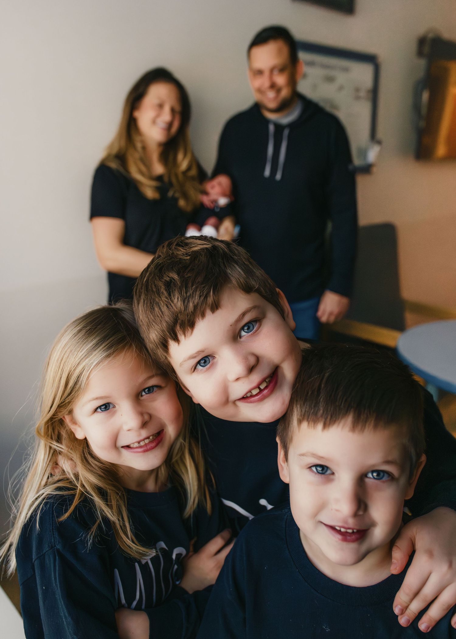 Three young children smile while posing together for a family photo with parents in the background.