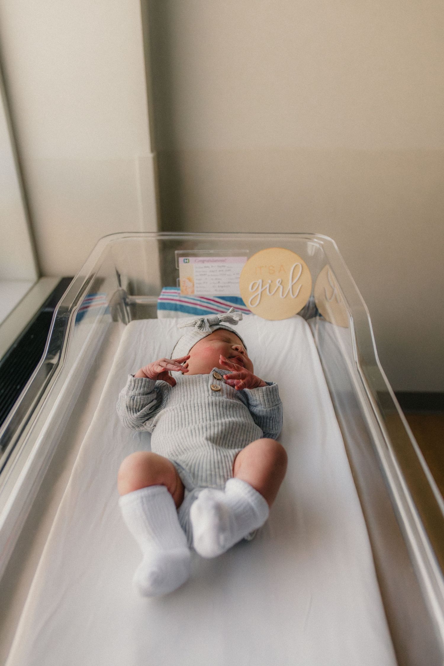 Newborn baby in gray outfit lies on white changing pad wearing white socks with a name tag visible above.