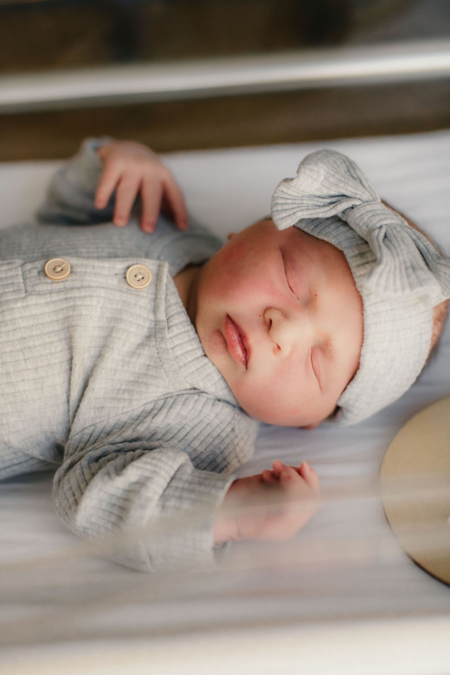 A sleeping newborn wearing a gray outfit and white bow headband rests peacefully.