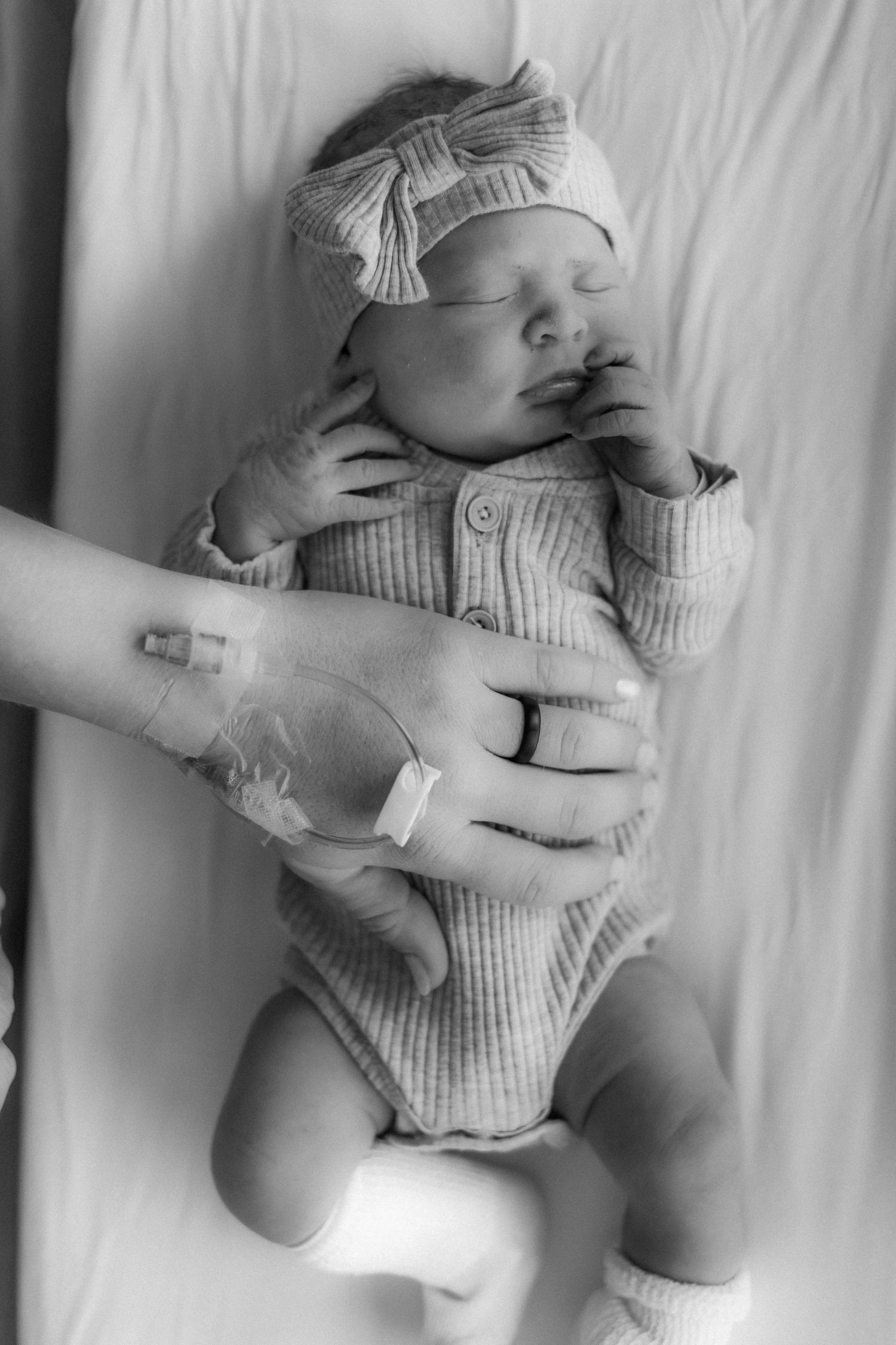A sleeping newborn baby wearing a bow headband and ribbed onesie lies on white bedding as adult hands gently touch them.