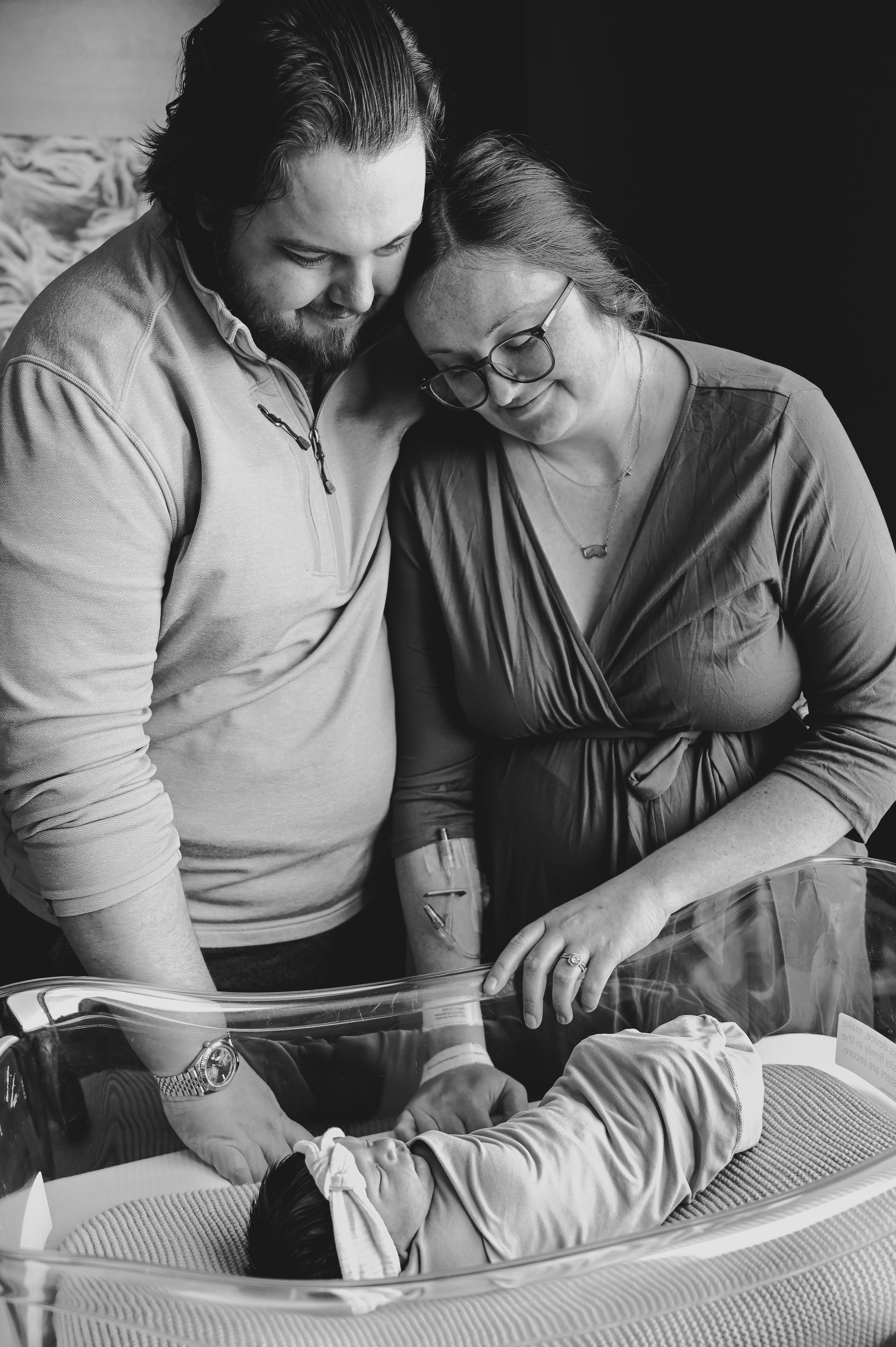 Black and white photo of new parents lovingly gazing at their newborn baby in a hospital bassinet.