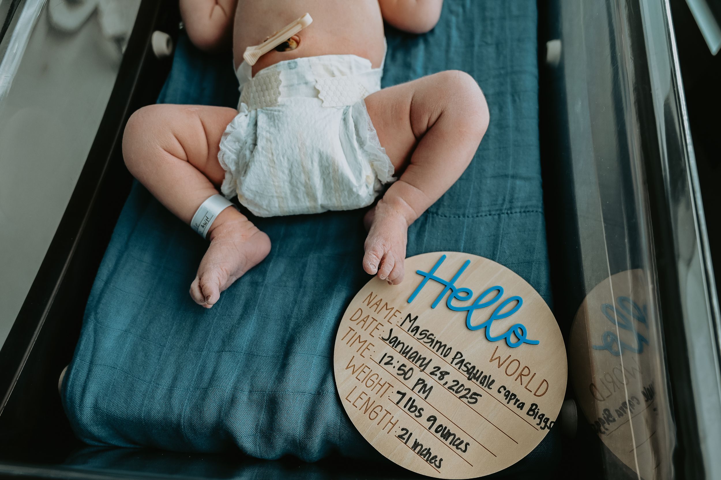 Newborn baby lying on blue blanket next to a Hello World birth announcement sign in hospital.
