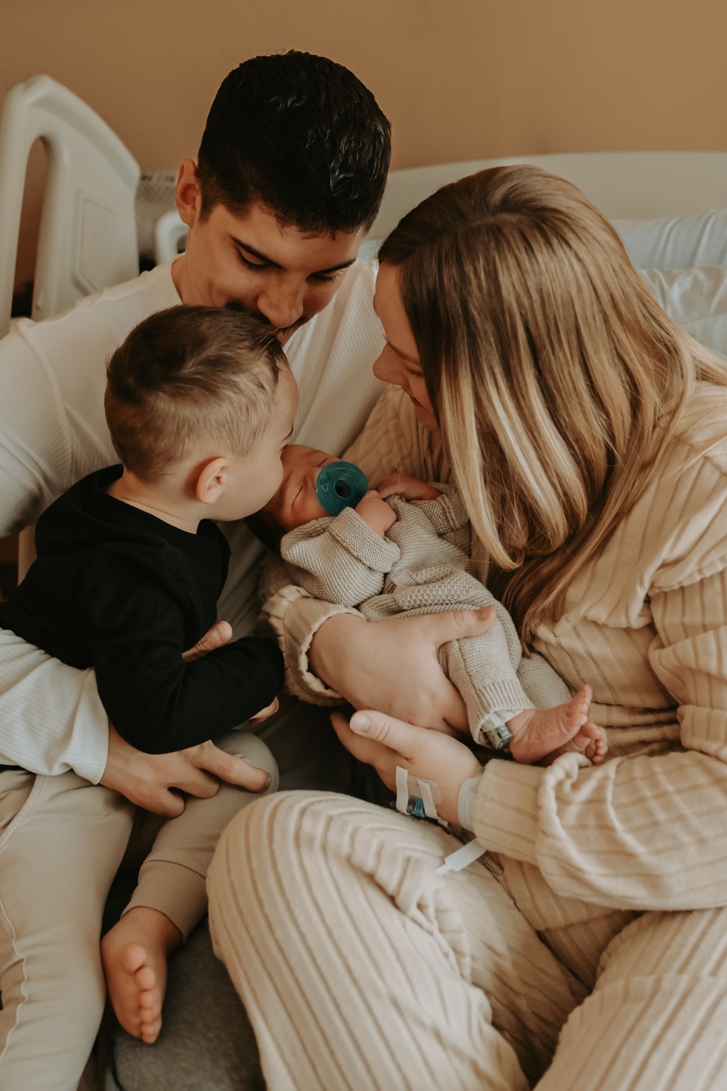 A young family with a toddler boy tenderly gazes at their newborn baby in a warm hospital room.