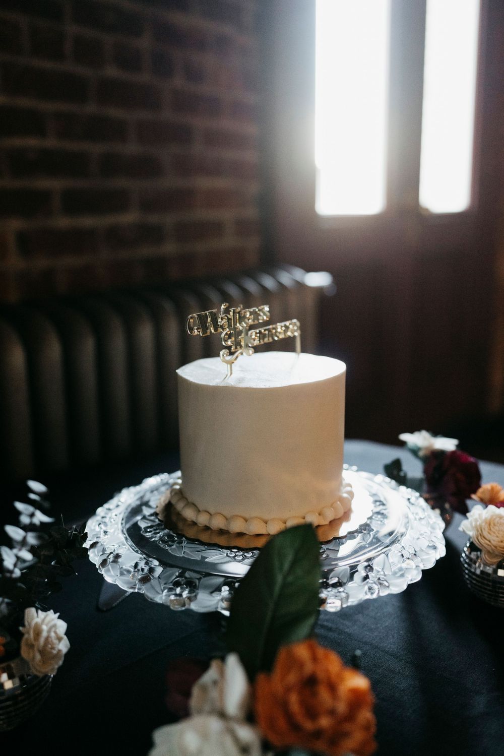 Elegant white wedding cake on crystal plate with gold crown topper and floral decorations against brick wall.
