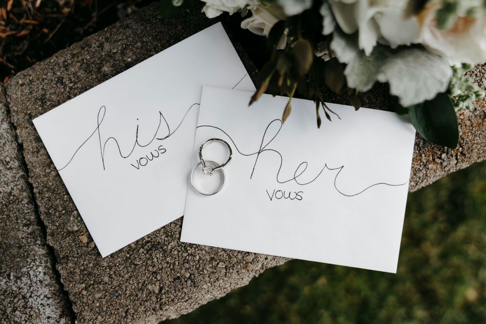 Wedding rings sit between two cards labeled 'his' and 'her' vows on a grey stone surface with white flowers in background.