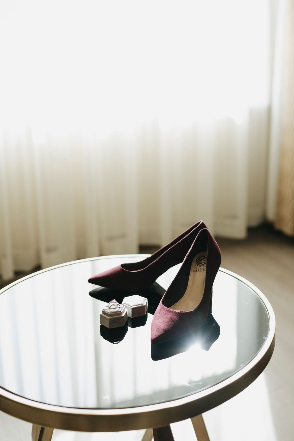 Elegant black high heels and wedding rings displayed on a round mirrored table with sheer curtains in background.