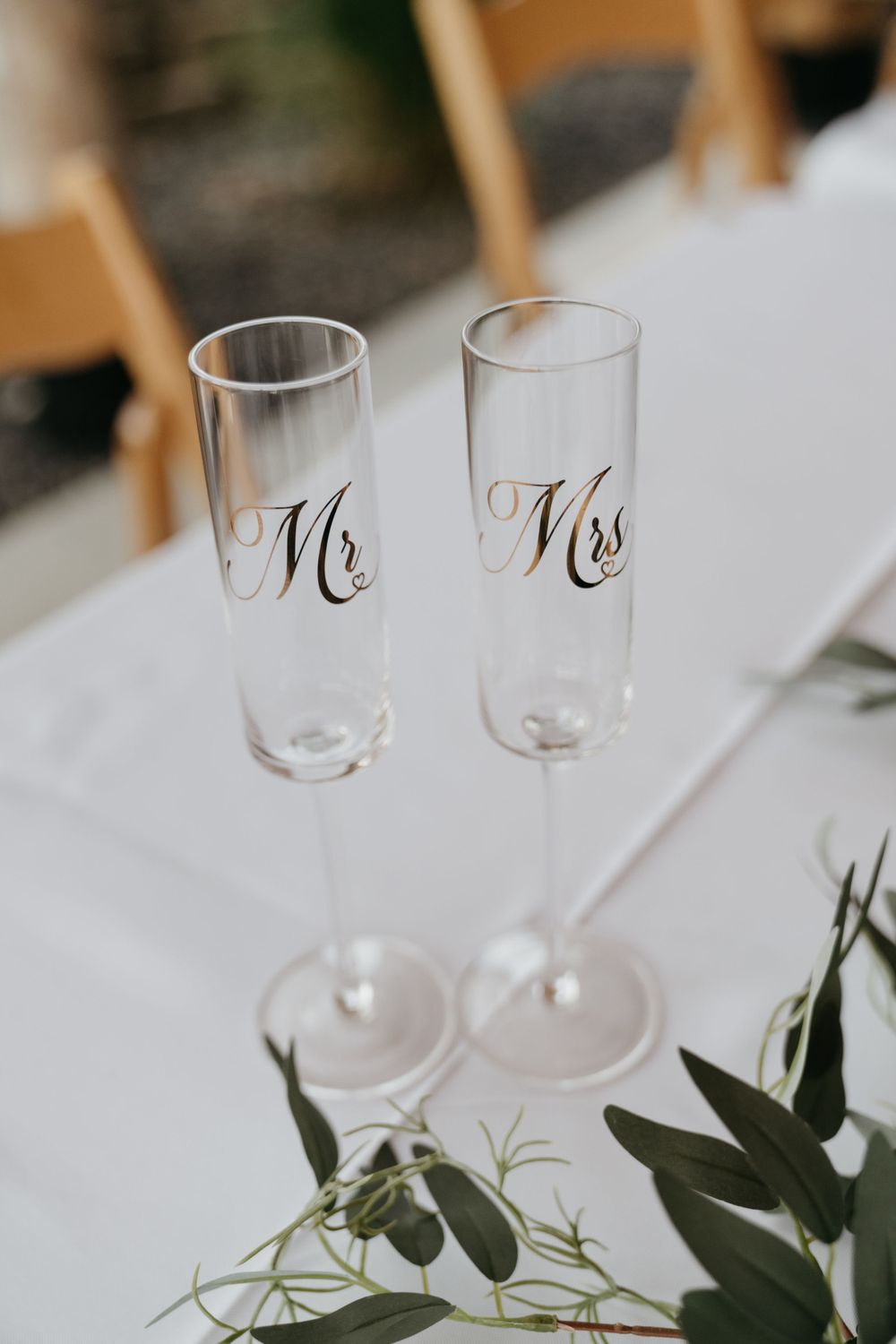 Two elegant champagne flutes with 'Mr' and 'Mrs' text placed on a white tablecloth with greenery decorations.