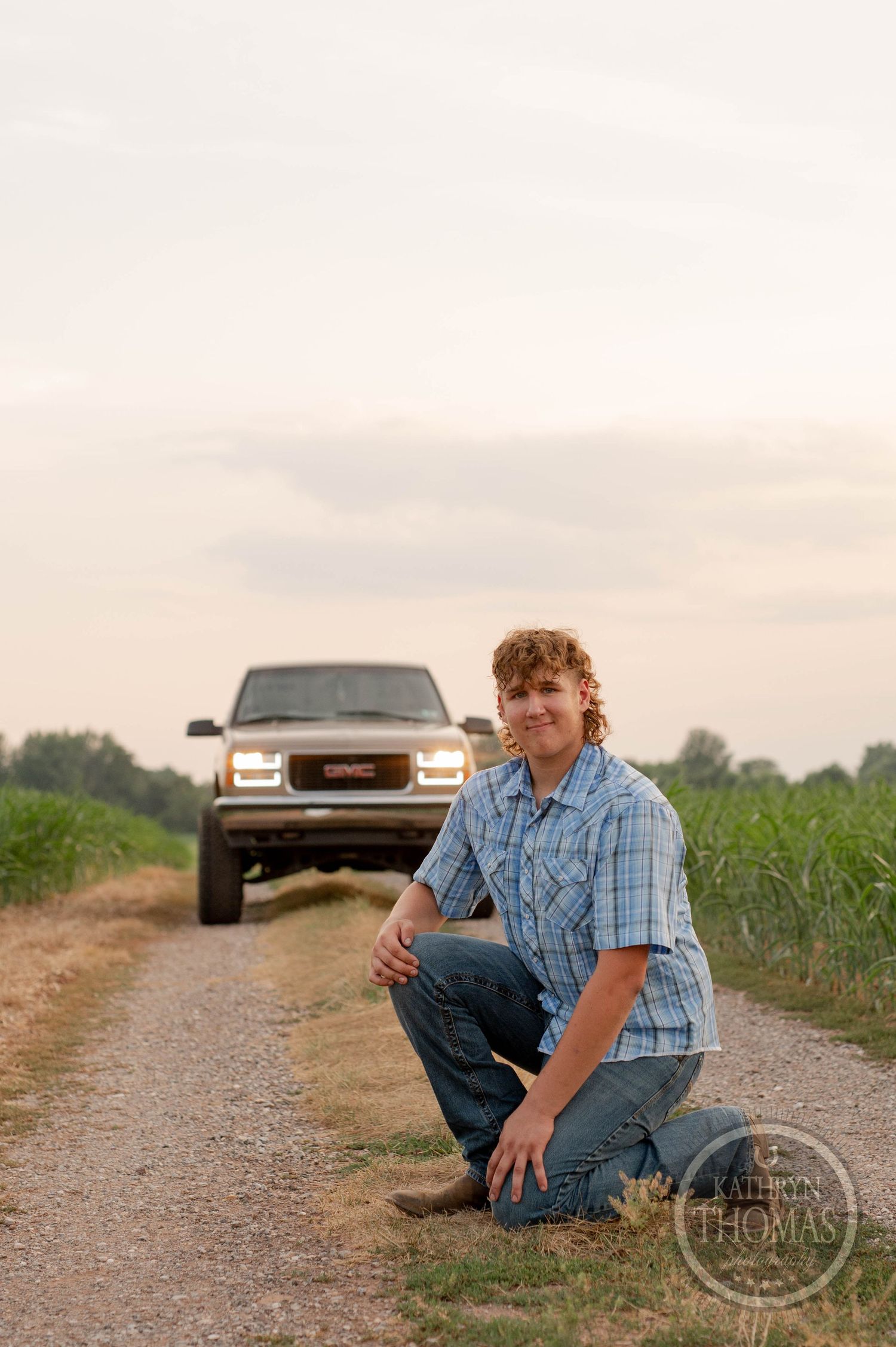 Person kneeling on dirt road with lifted GMC truck parked in background during sunset.