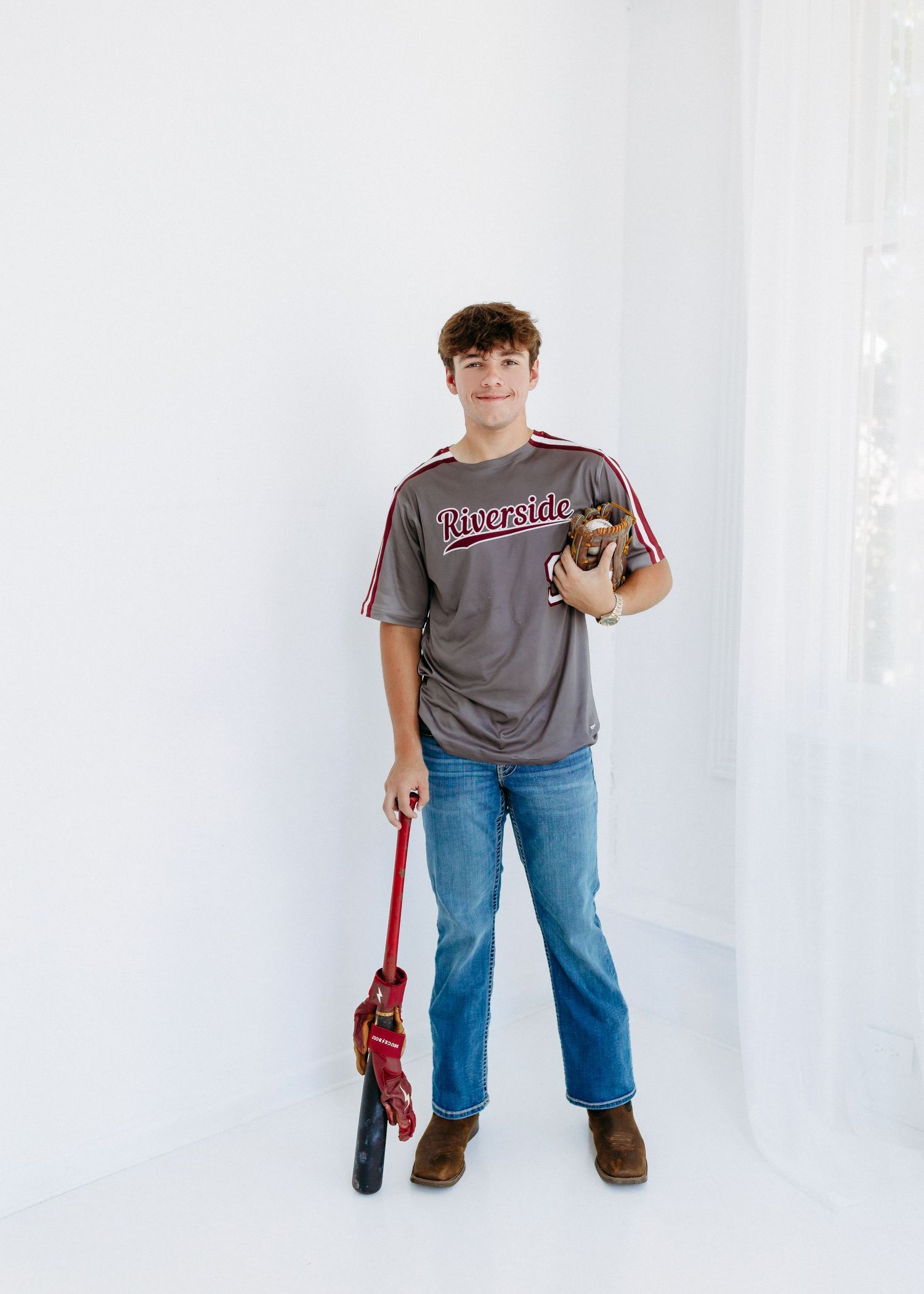Student in casual attire holding football and red baseball bat poses against white backdrop in studio setting.