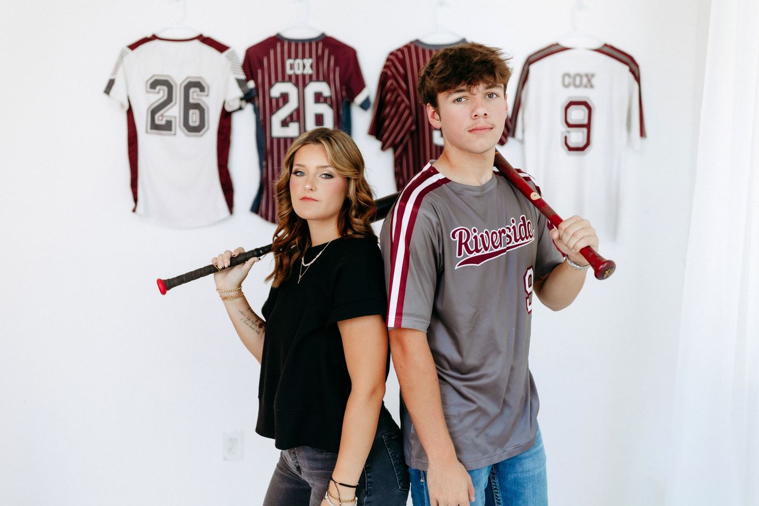 Two young baseball players stand back-to-back holding bats with sports jerseys displayed on the wall behind them.