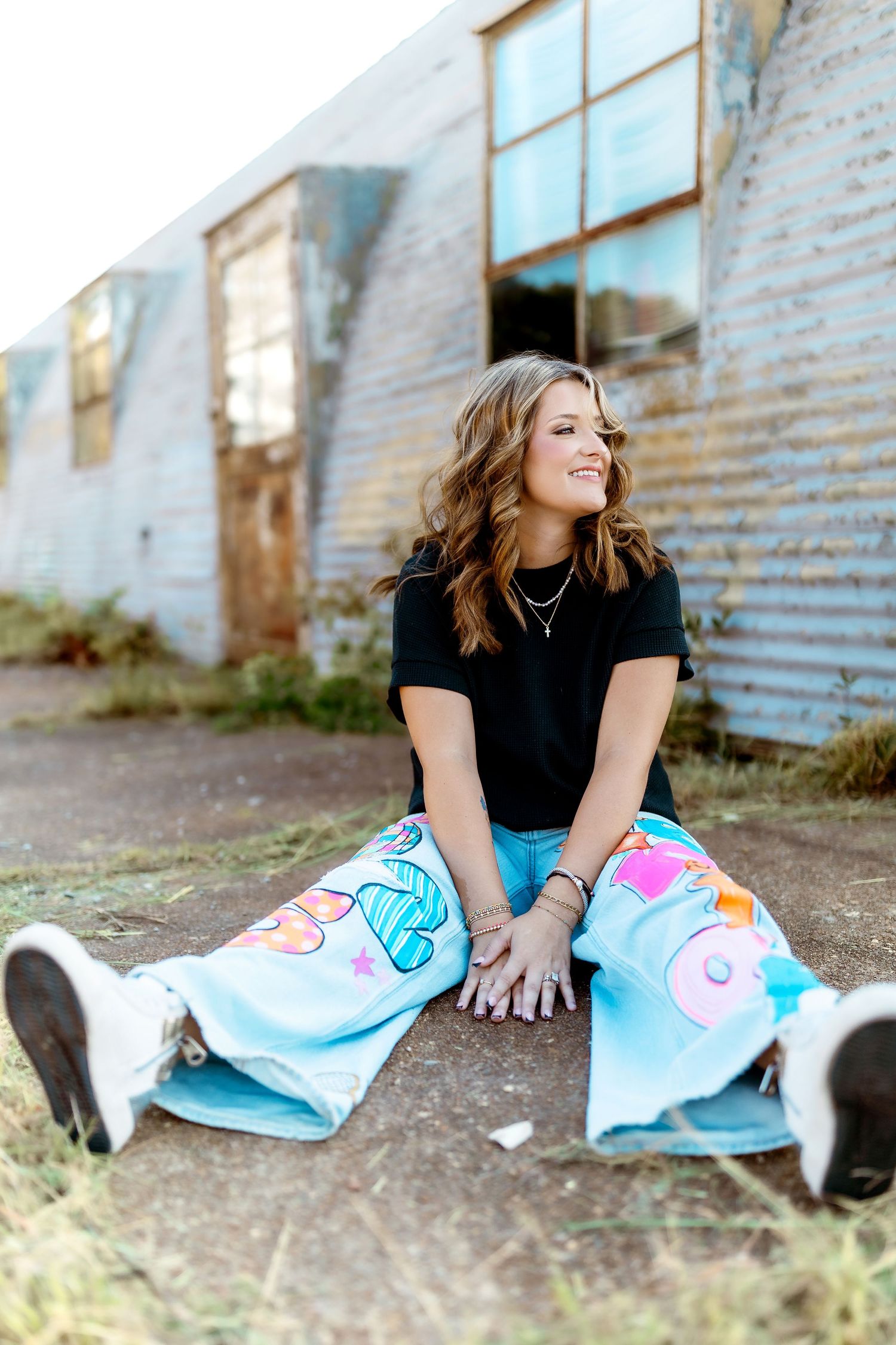 Woman in black shirt and colorful painted jeans sits casually against rustic metal wall during outdoor portrait session.