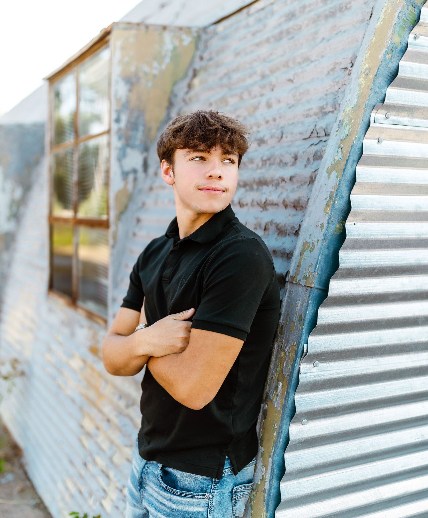 Young person in a black polo shirt and jeans poses with arms crossed against a corrugated metal and brick wall exterior.