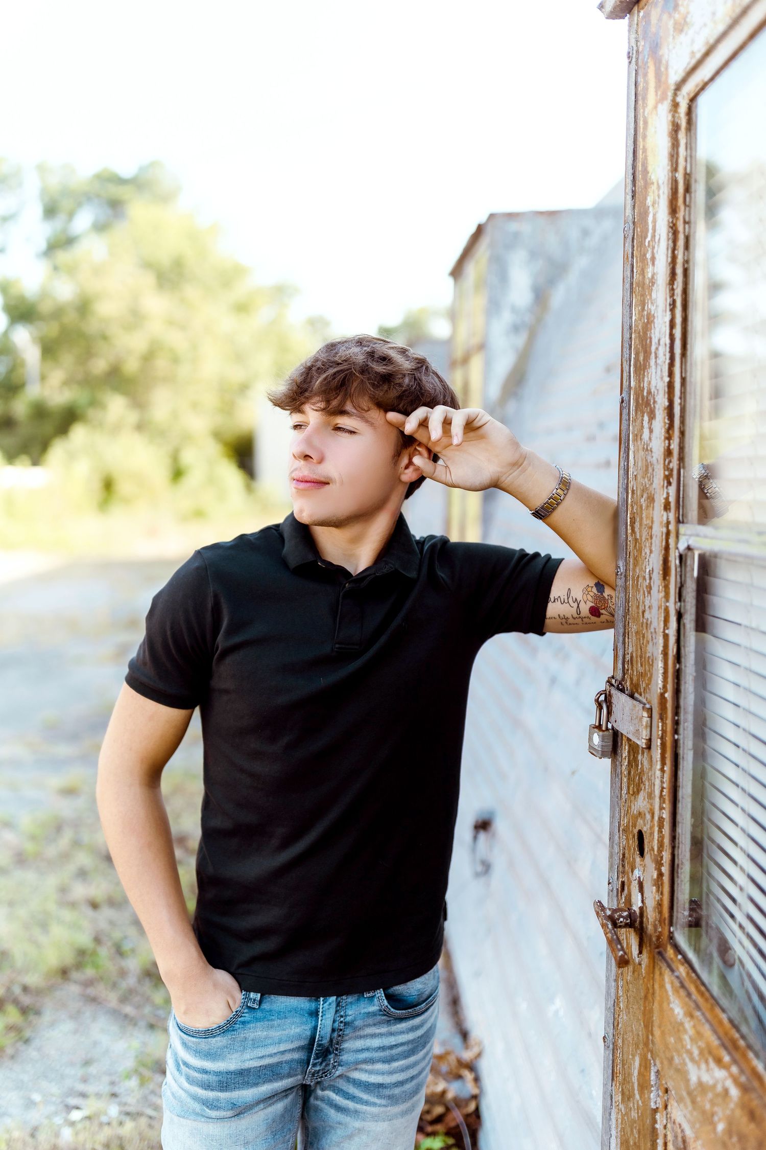 Person in a black polo shirt and jeans poses casually against a rustic white wall in warm outdoor lighting.