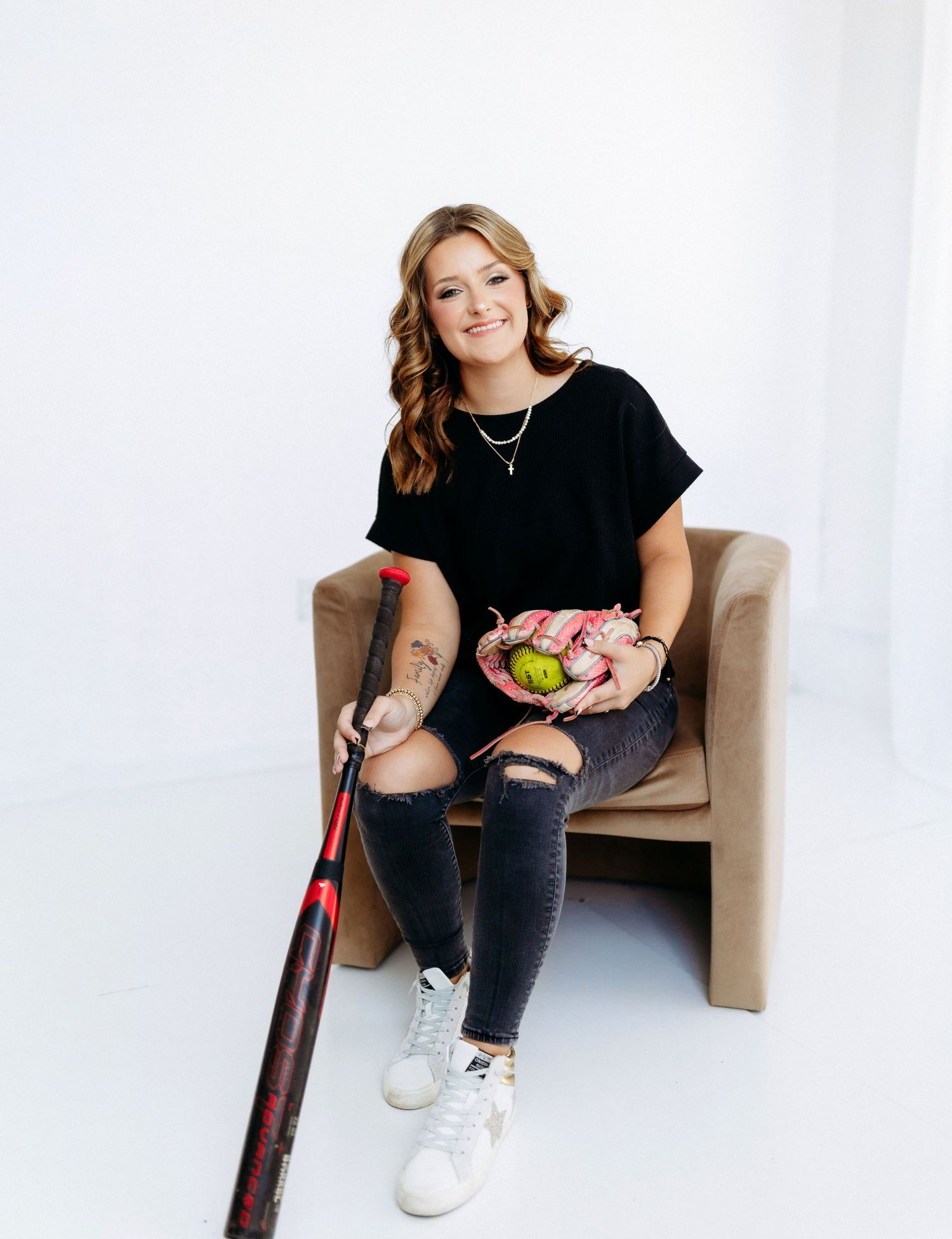 A person in black attire sits on a brown chair holding a baseball bat and colorful flowers against a white backdrop.