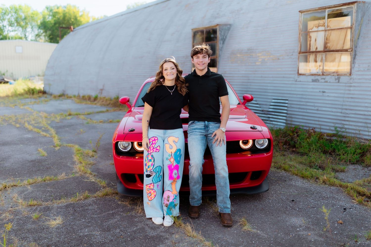 A bright red Dodge Challenger parked outside a corrugated metal building with two people standing in front.