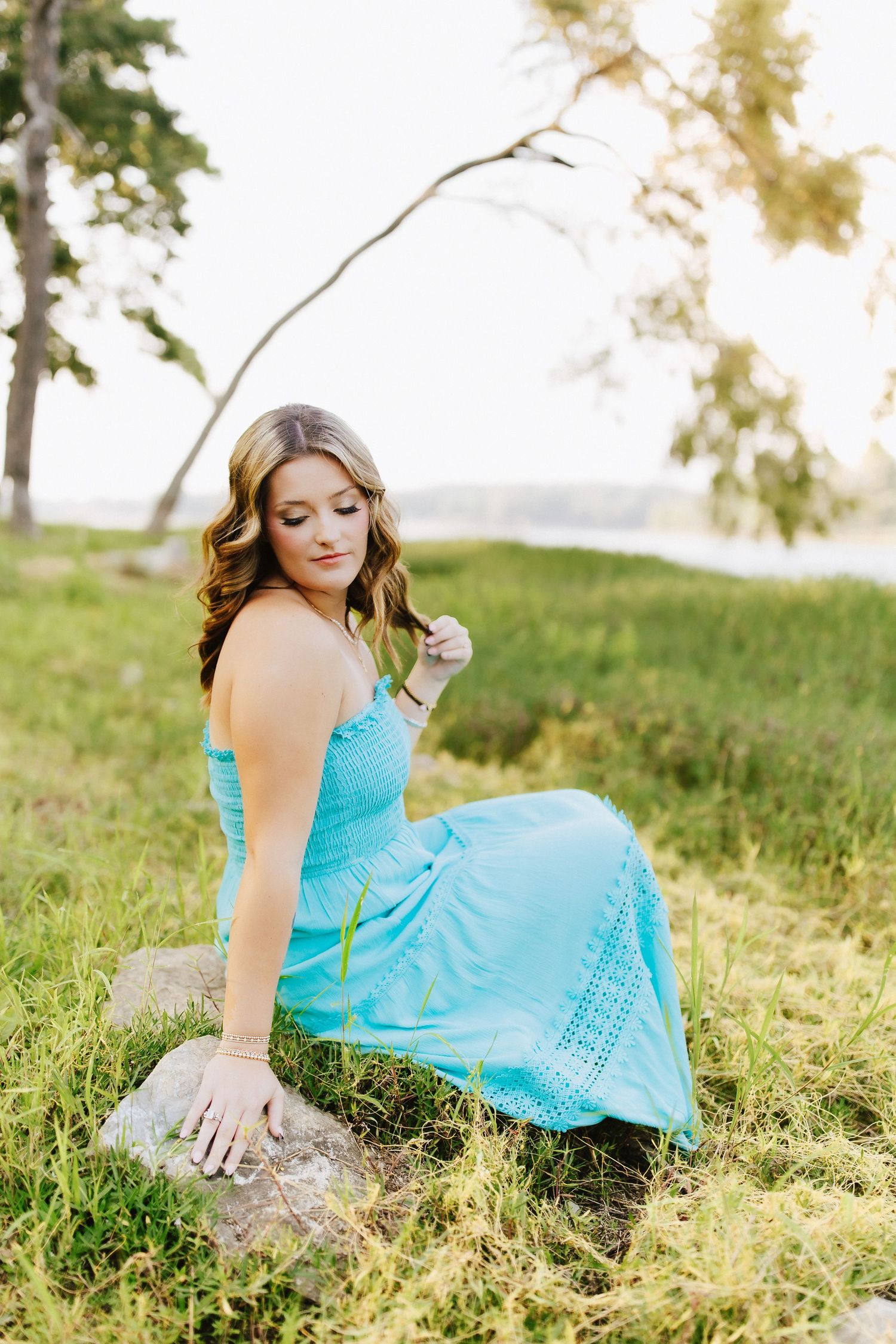 In a turquoise lace dress, a figure sits gracefully on a rock amid green grass during golden hour.