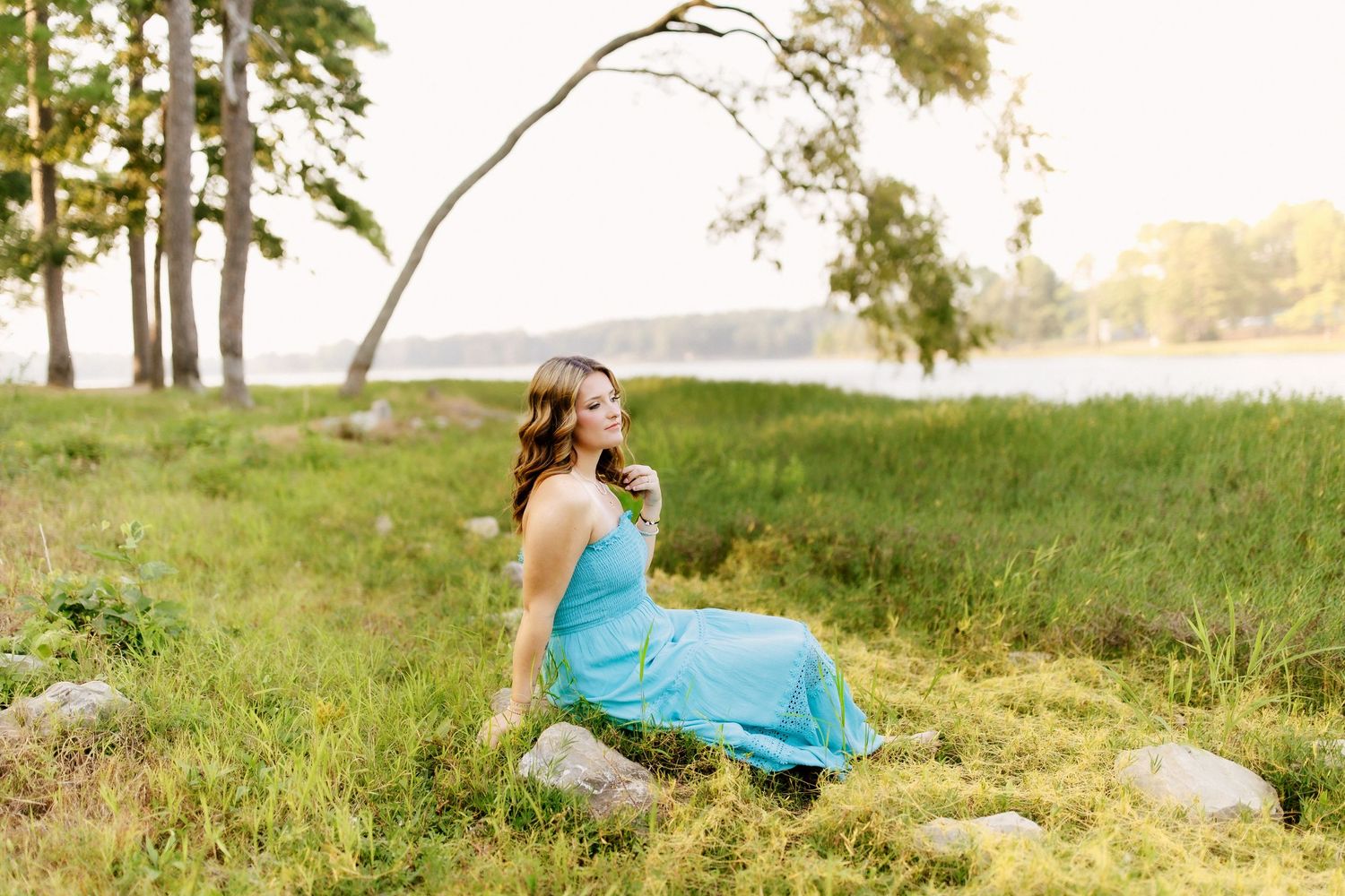 Someone in a turquoise dress sits on a rock by a lakeside during golden hour with trees in the background.