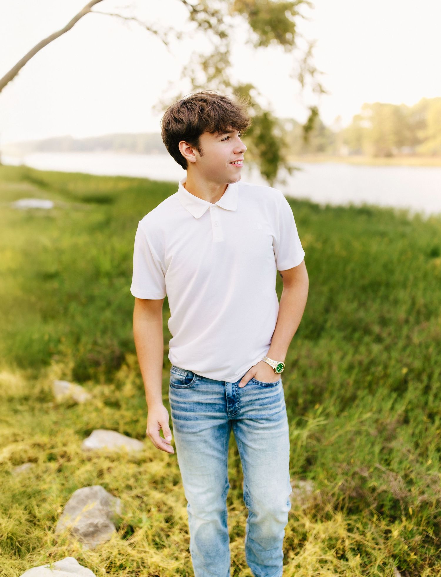 Person in a white polo shirt and light wash jeans poses casually outdoors in natural sunlight by grass and water.