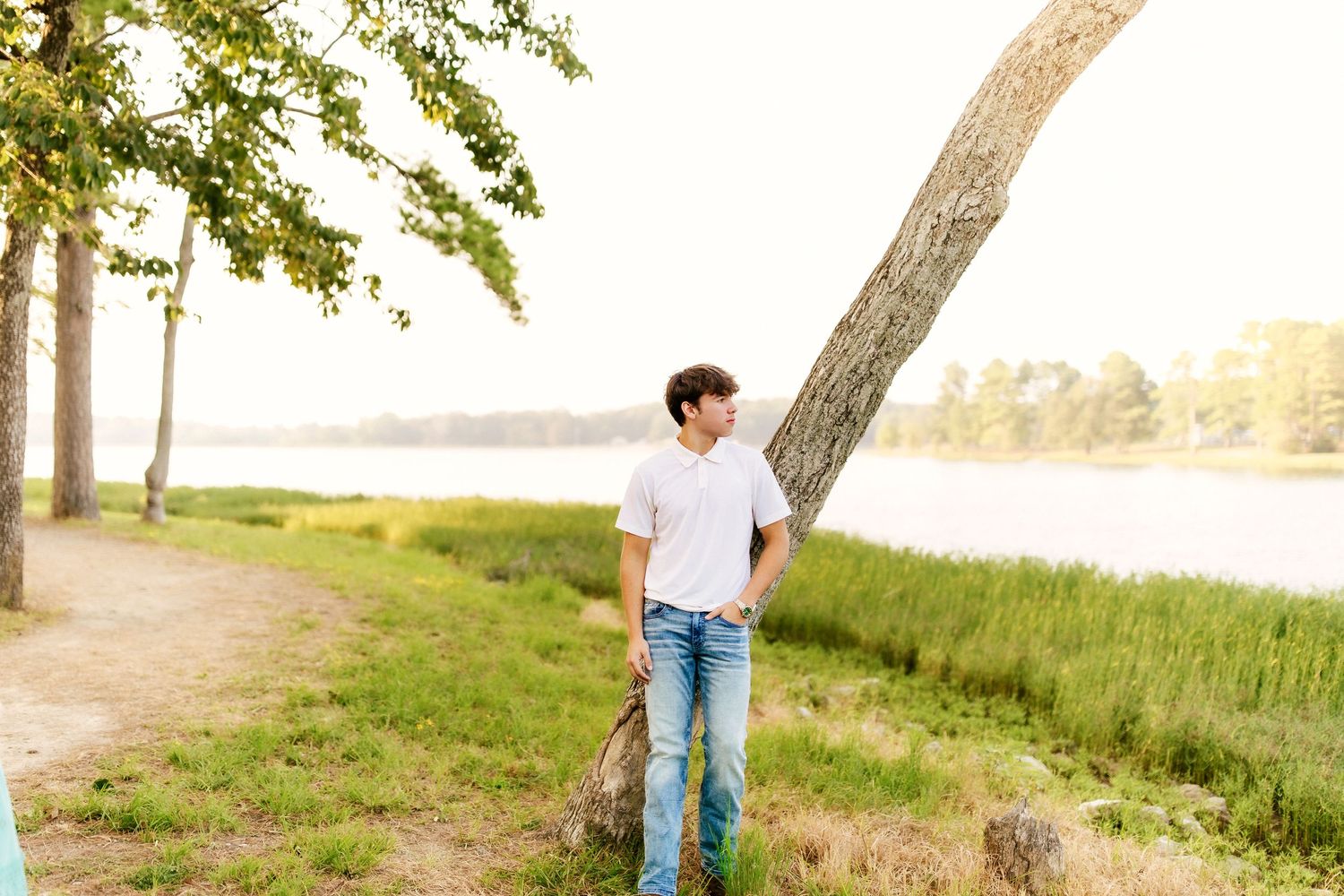 A person in casual wear stands near a leaning tree beside a tranquil lake during golden hour.