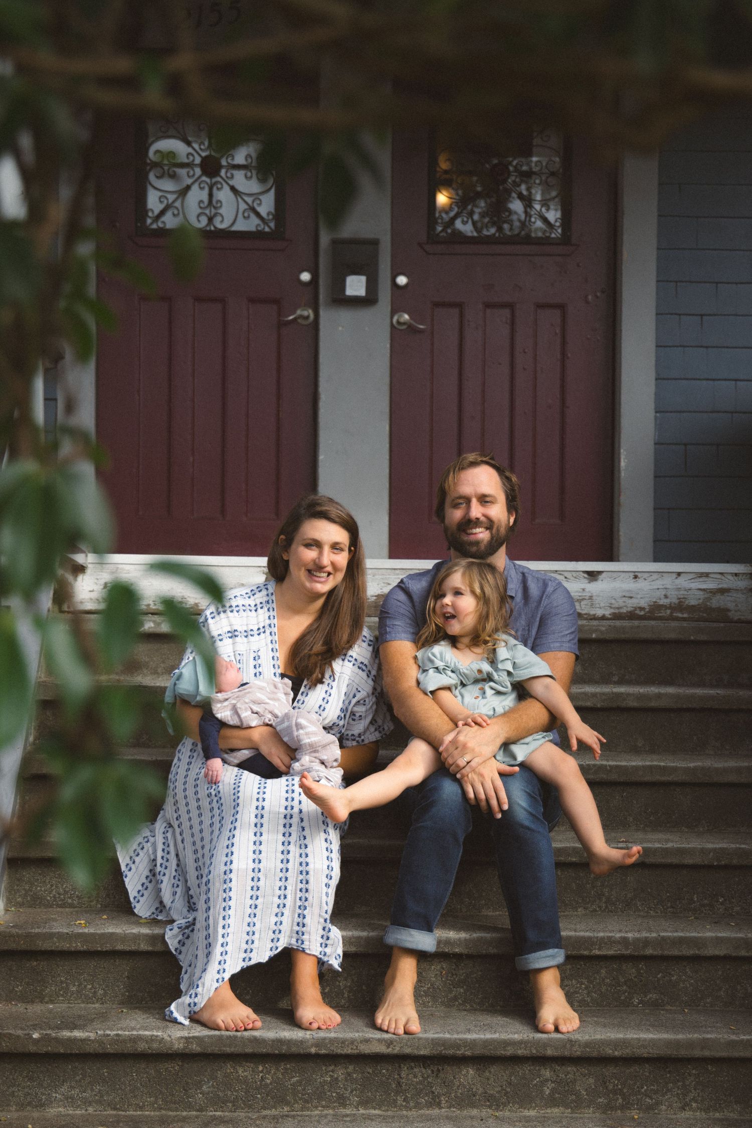 Family of four with newborn and toddler posing on front steps of their house for a family photo