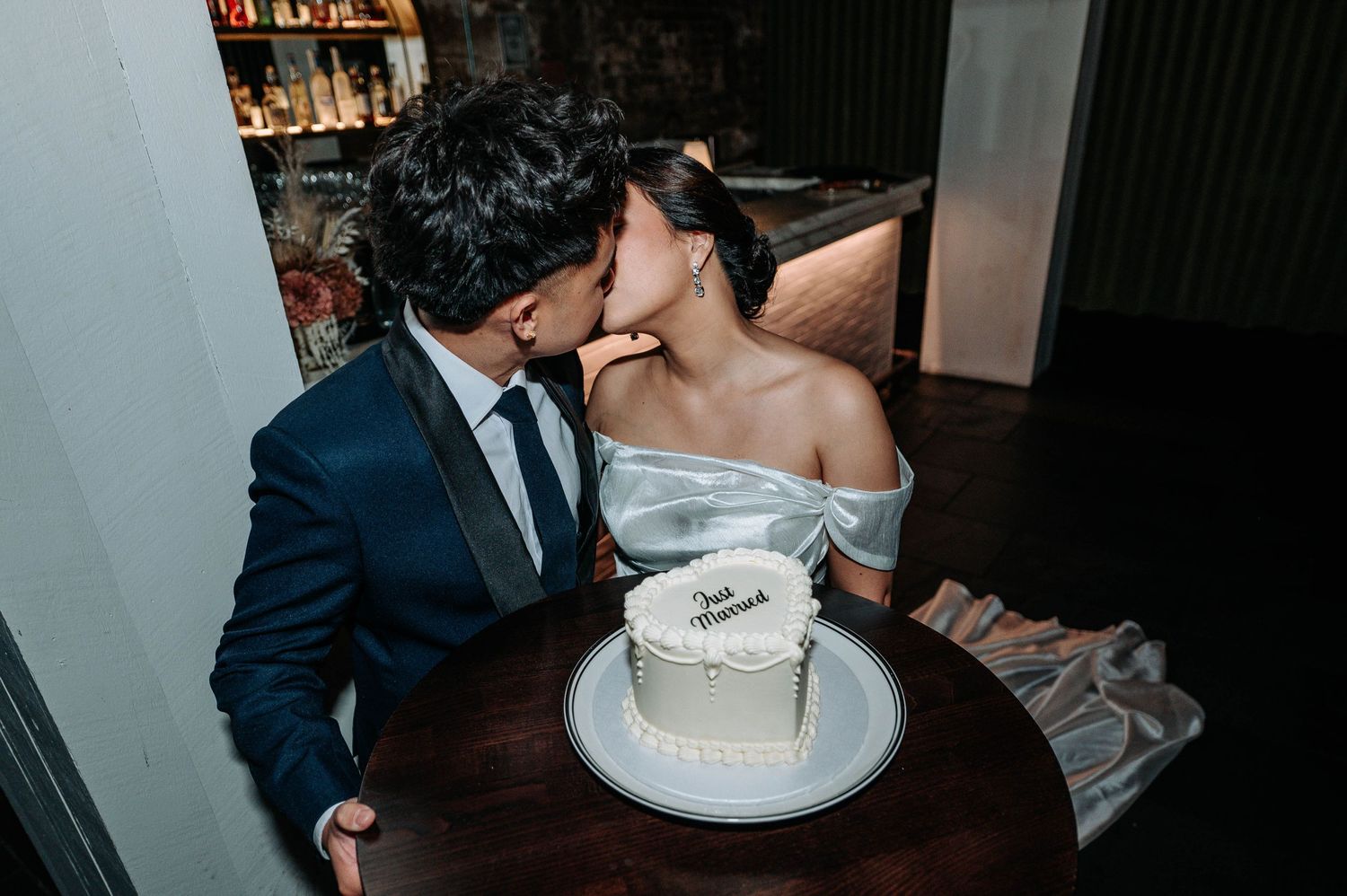 A couple shares an intimate moment over a small white wedding cake at a bar.