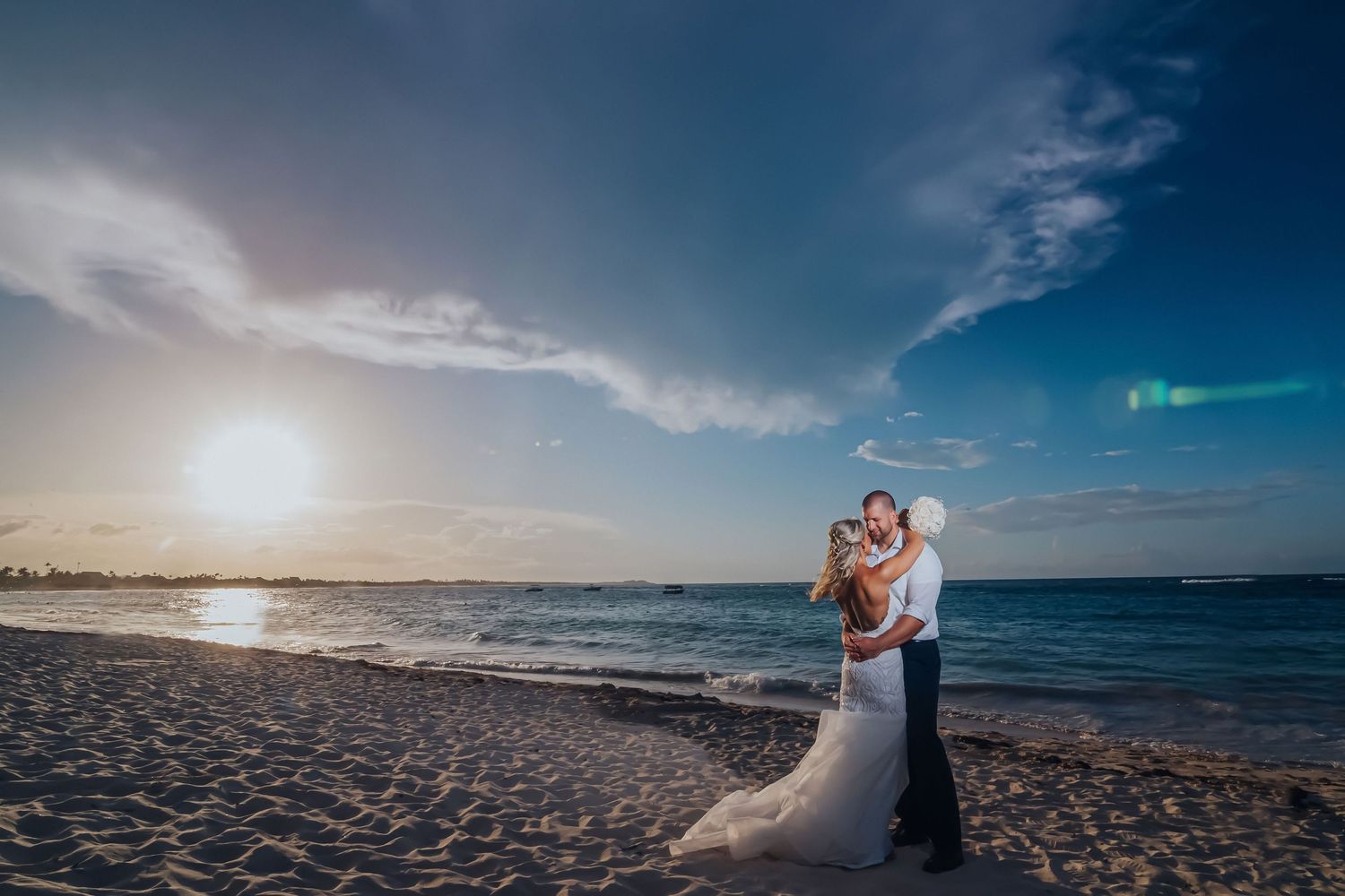 Couple embracing on beach at sunset with dramatic sky and ocean backdrop.
