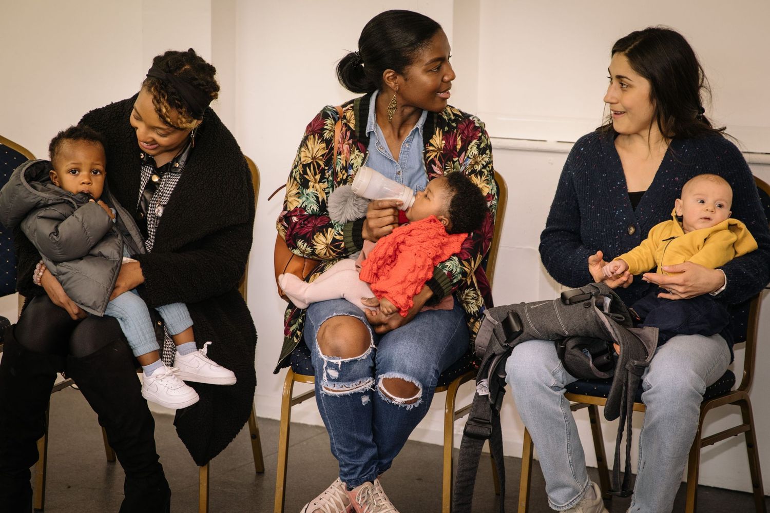 A group of adults sitting in chairs cradling and caring for young infants in a waiting room setting.