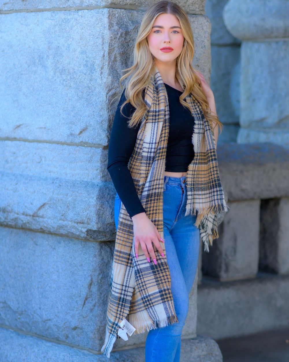 Casual winter fashion look featuring a plaid scarf, black top, and blue jeans against a stone wall backdrop.